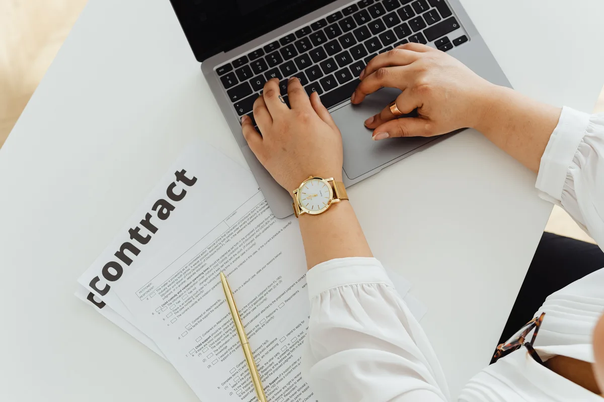 A person working on a laptop beside a printed contract, reviewing the statement of work for AI agent development services