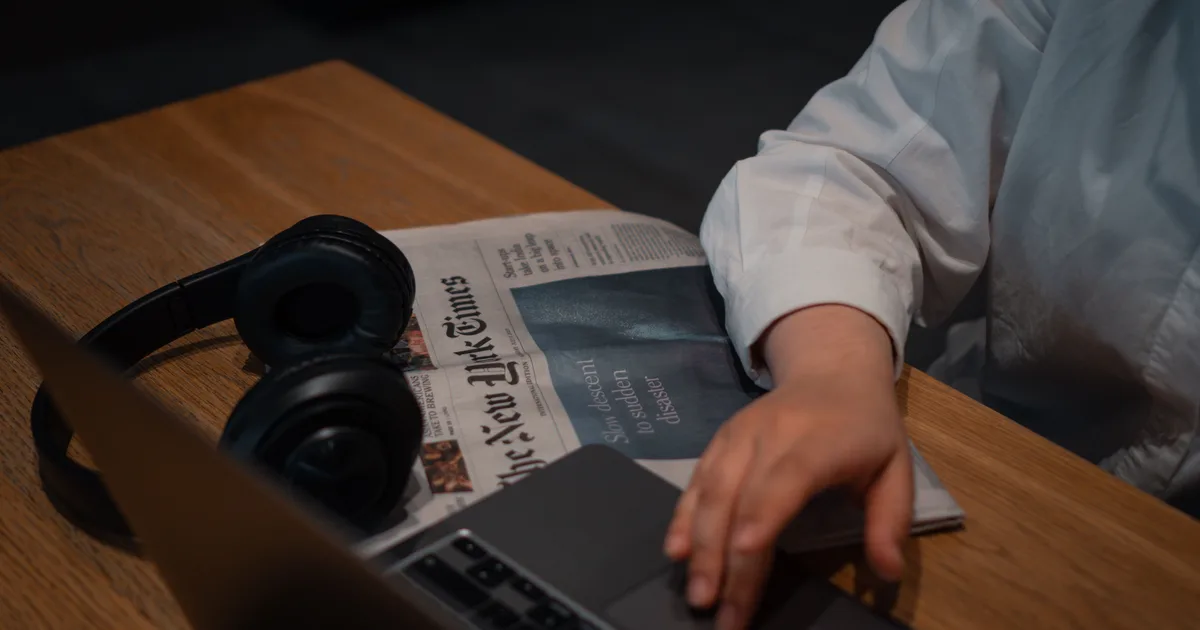 A person working on a laptop beside a newspaper and headphones, catching up on AI agents news in 2026