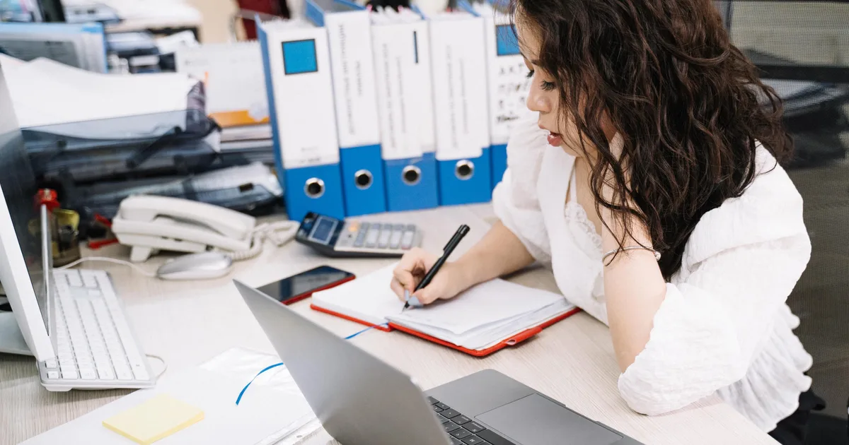 Caseworker taking notes at an office desk beside a laptop and a row of policy binders, the setting an AI caseworker policy assistant is built for
