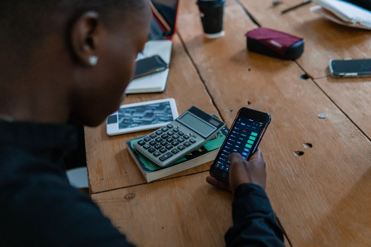 A person checking a finance app on a smartphone at a wooden desk with a calculator beside them, the kind of task people now hand to an AI personal assistant