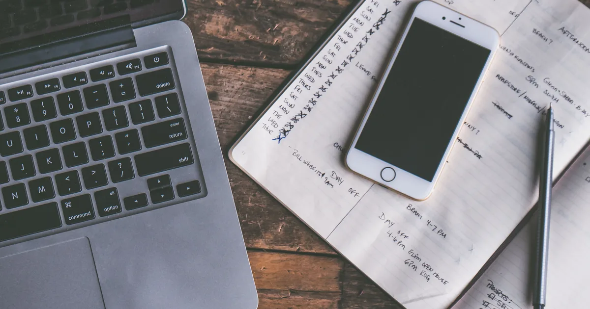 A laptop, a smartphone and an open handwritten planner on a wooden desk, the daily schedule an AI personal assistant is meant to manage
