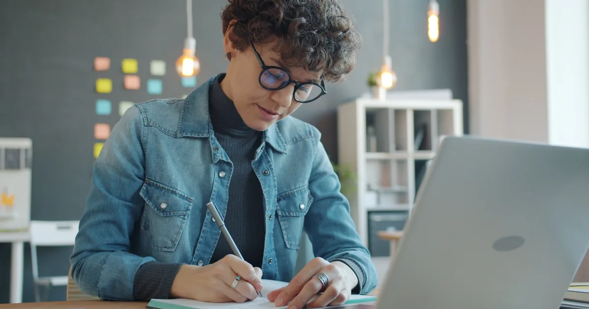 Woman writing notes at a desk beside her laptop, planning a job search for AI prompt engineer jobs