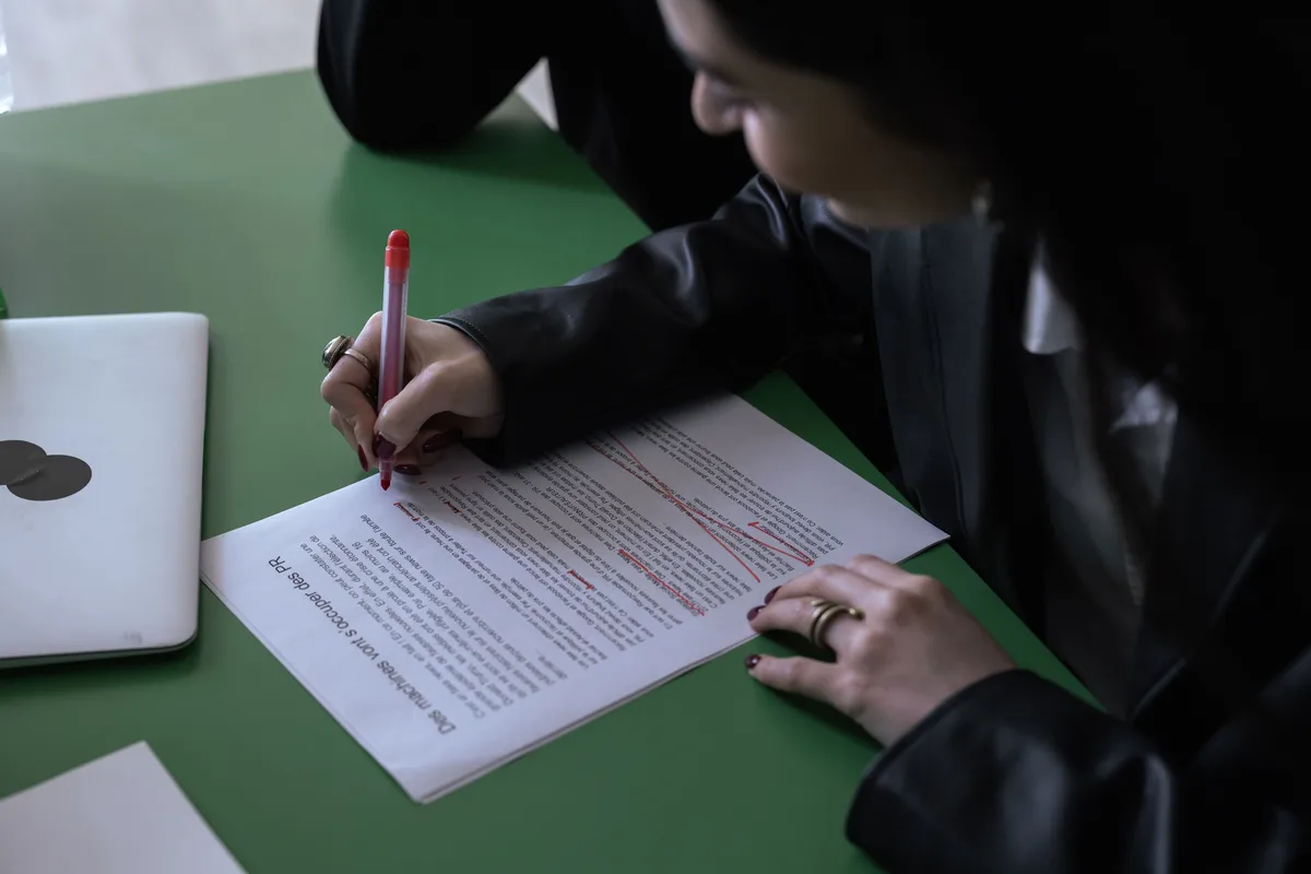 A person marking up a printed document in red pen next to a laptop, the editing pass that no Claude prompt to bypass AI detection can replace