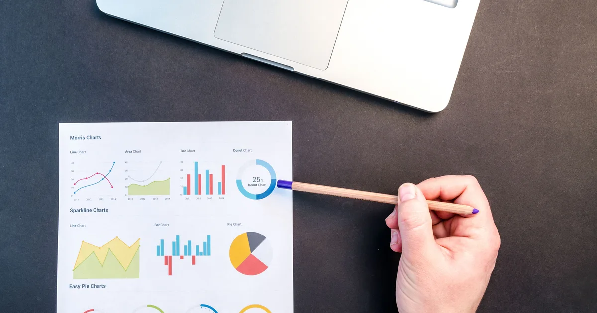 Hand with a pencil reviewing printed financial charts beside a laptop, the kind of analysis explainable AI in finance has to make traceable