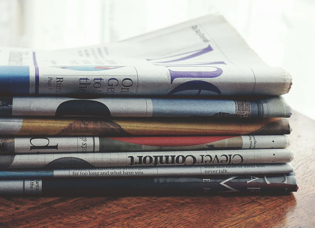 A stack of folded newspapers on a wooden table, the kind of news coverage AI assistants summarise when you ask for today's headlines