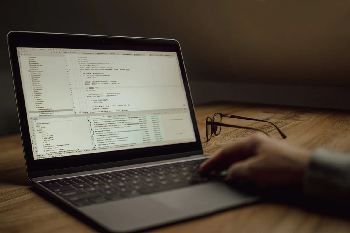 A developer working on code on a laptop at a desk with glasses beside the keyboard, reviewing changes from a text editor AI agent