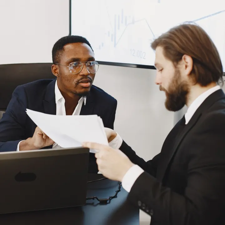 A consultant and a client discussing documents across a desk with a laptop